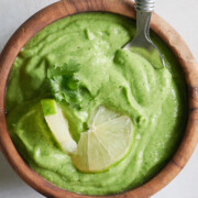 Wooden bowl of avocado green goddess dressing with lime pieces and cilantro on top sitting on white background.