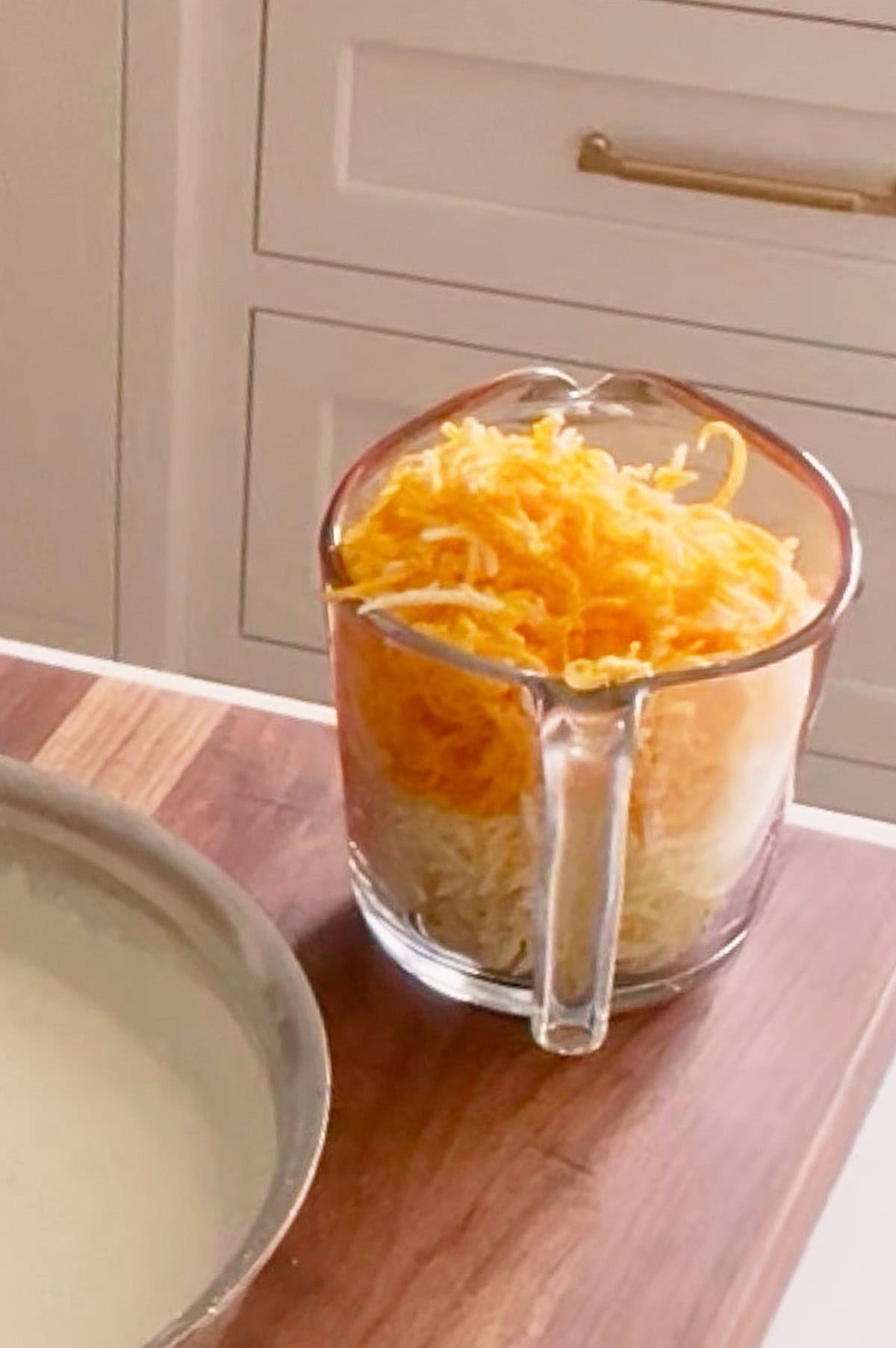 A glass measuring cup filled with shredded yellow and white cheese for four cheese mac and cheese sits on a wooden countertop next to a round dish.