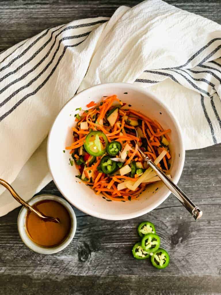 white bowl with grated carrots, diced apples and jalapenos with dressing and a white and black striped dish towel on a gray wooden background.