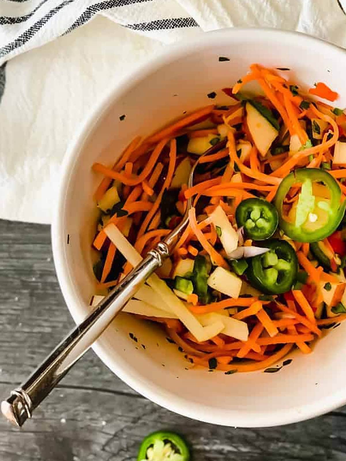 A white bowl filled with colorful slaw—julienned carrots, sliced jalapeños, cucumber, and apple strips—all mixed together with a fork resting inside.