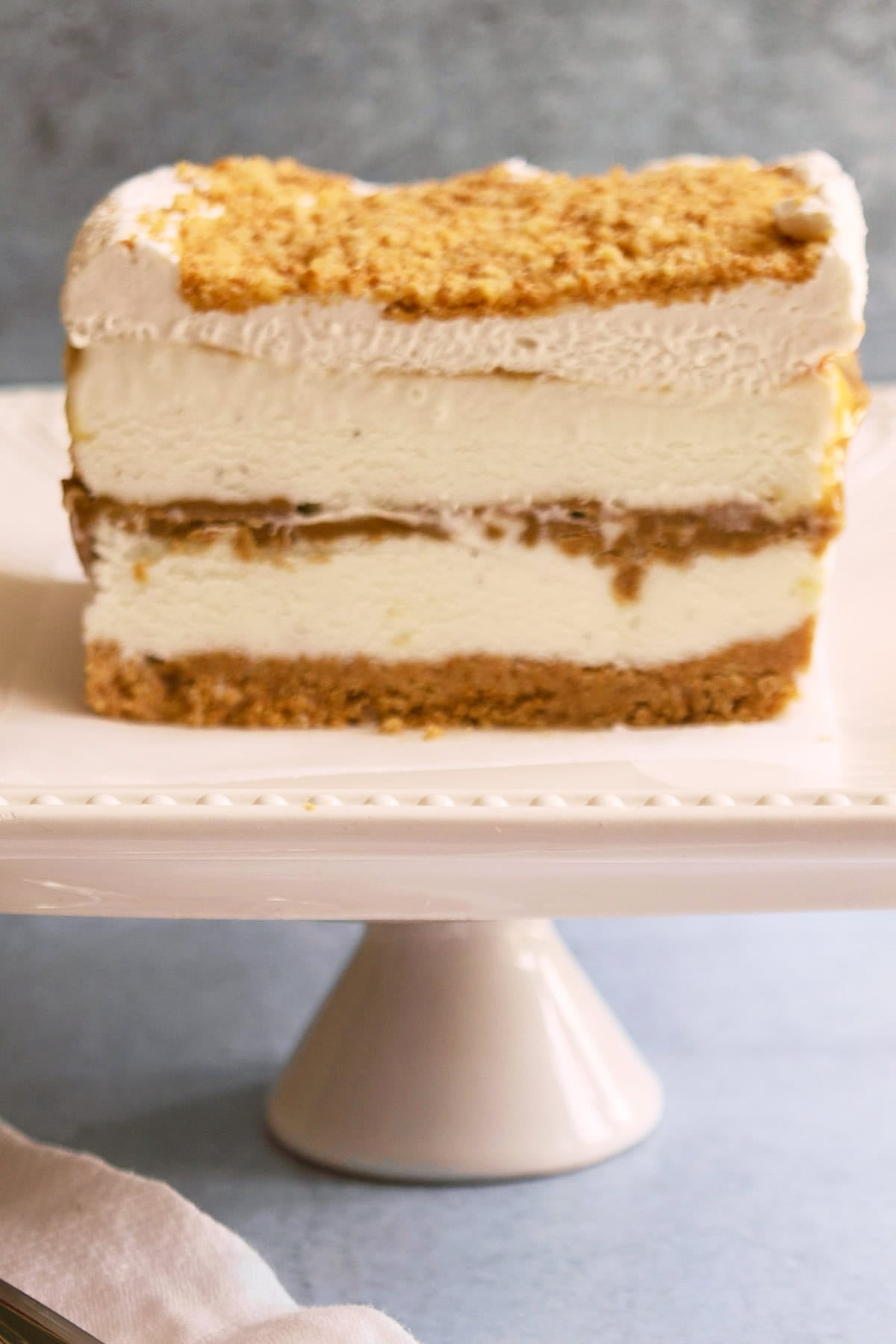 A slice of layered ice cream cake with a cookie crust, ice cream filling, a middle layer of Biscoff, and a crumb topping on a pedestal plate.