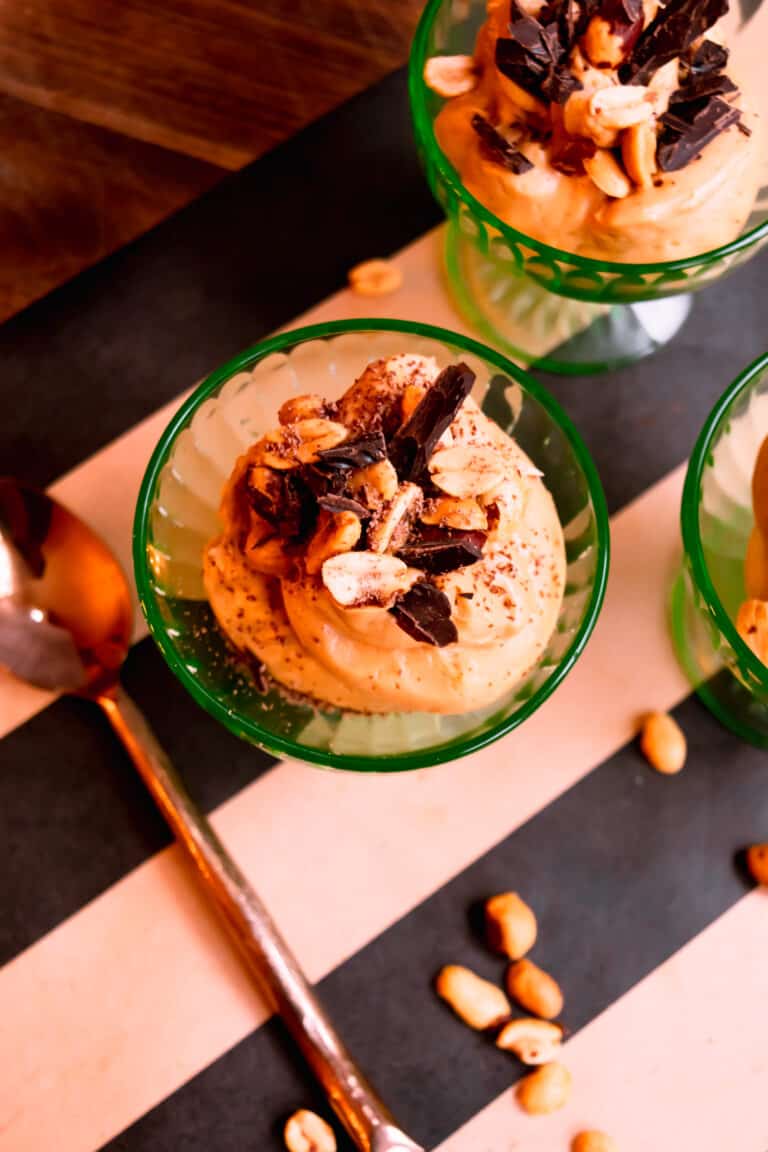 close up of a green bowl of peanut butter mousse topped with chopped chocolate and peanuts on a black and brown striped runner with a copper spoon.