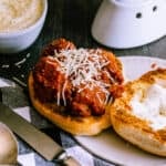 meatball sandwich on a roll with a black and white gingham napkin, silverware and a bowl of parmesan on a dark background.