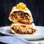 close up of two burritos stacked on top of each other with pickled jalapenos on top all on a white plate and some cilantro.