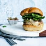 turkey burger on a white marble board with utensils and a bowl of caramelized onions to the side.