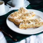 three pieces of Danish puff pastry on a white plate with two cups of coffee in the background on a blackwatch plaid napkin and white runner with a black backgrouund.
