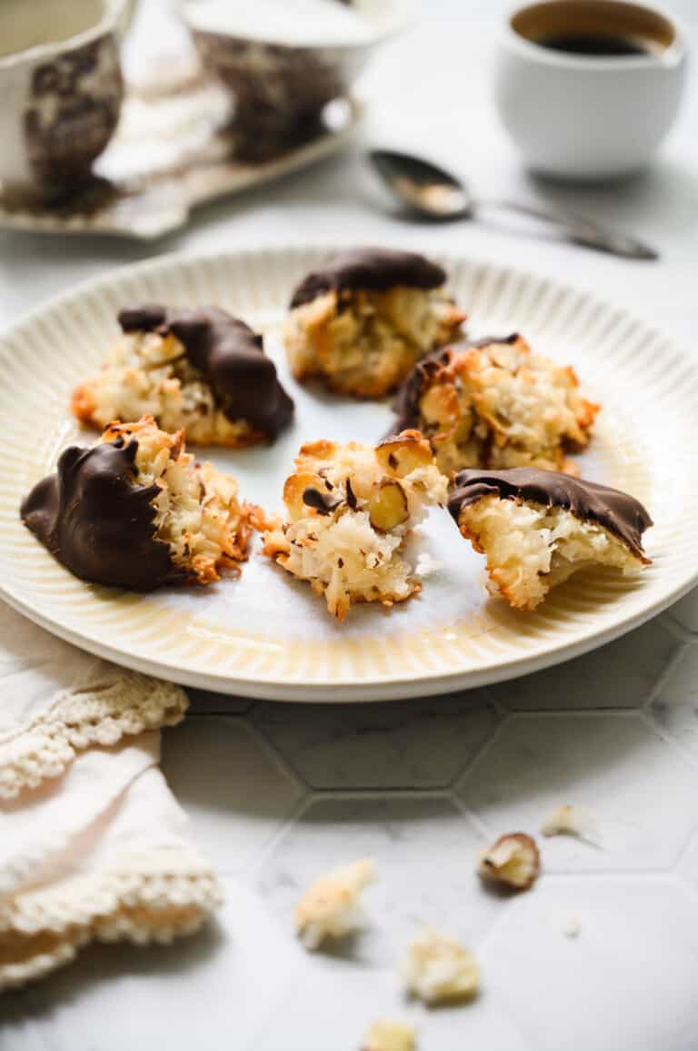 six coconut macaroons some dipped with chocolate on a white plate and silver spoon and cup of coffee