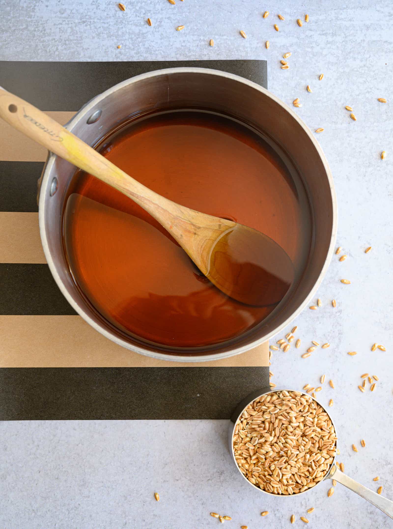 broth in a saucepan with a wooden spoon and a cup of farro in a measuring cup on a black and khaki striped runner.