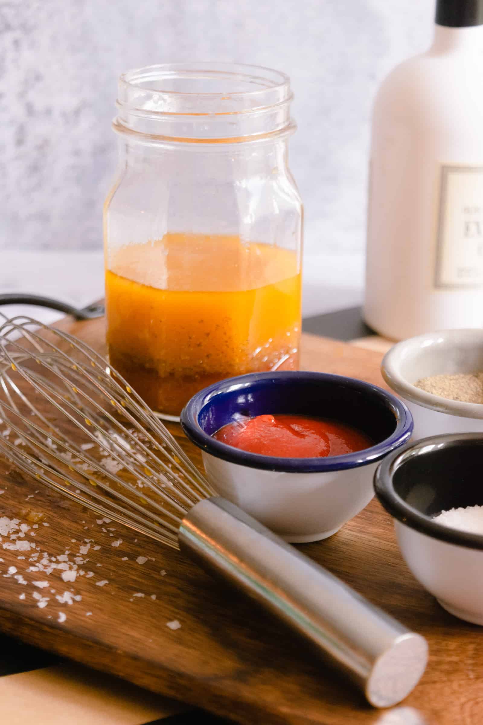 cutting board with a mason jar with salad dressing along with a whisk, sriracha, olive oil bottle and salt.