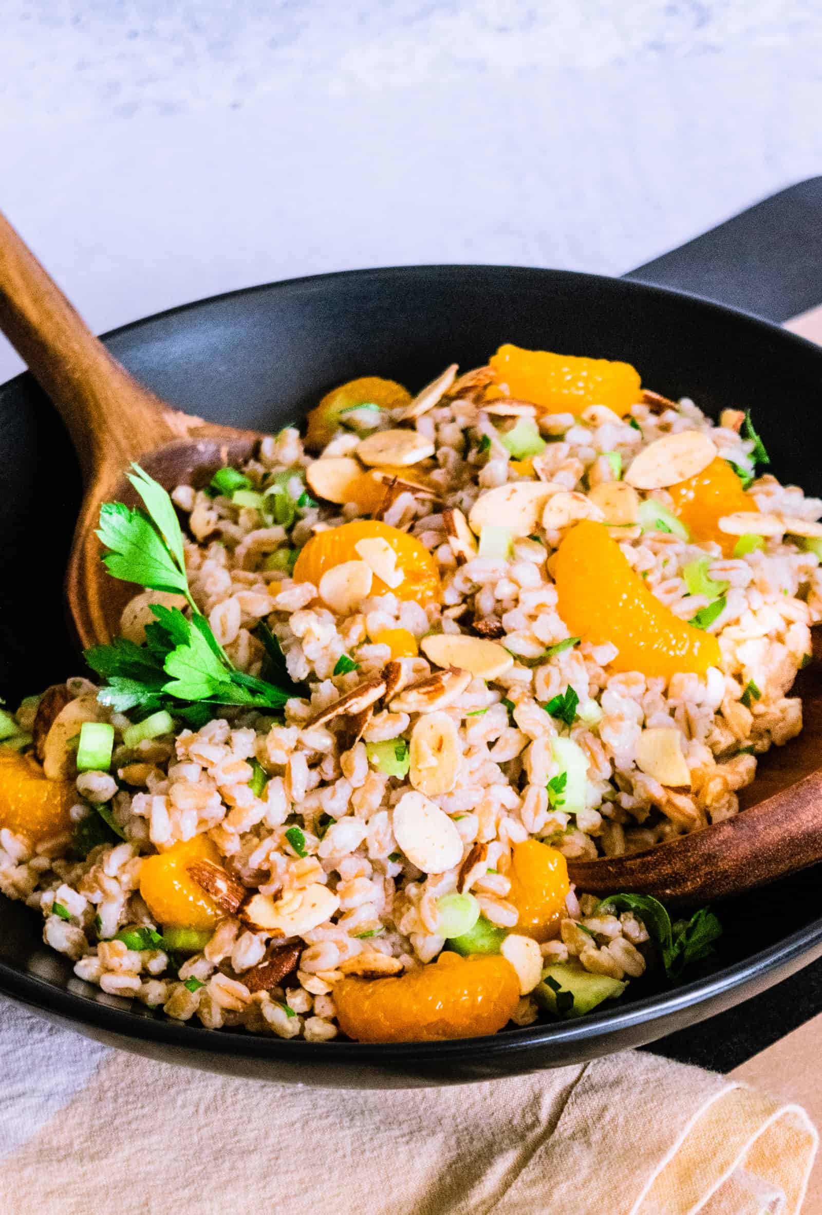 a black bowl with farro and orange salad with two wooden spoons on a black and khaki striped background.