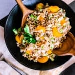 a black bowl with farro and orange salad with two wooden spoons on a black and khaki striped background