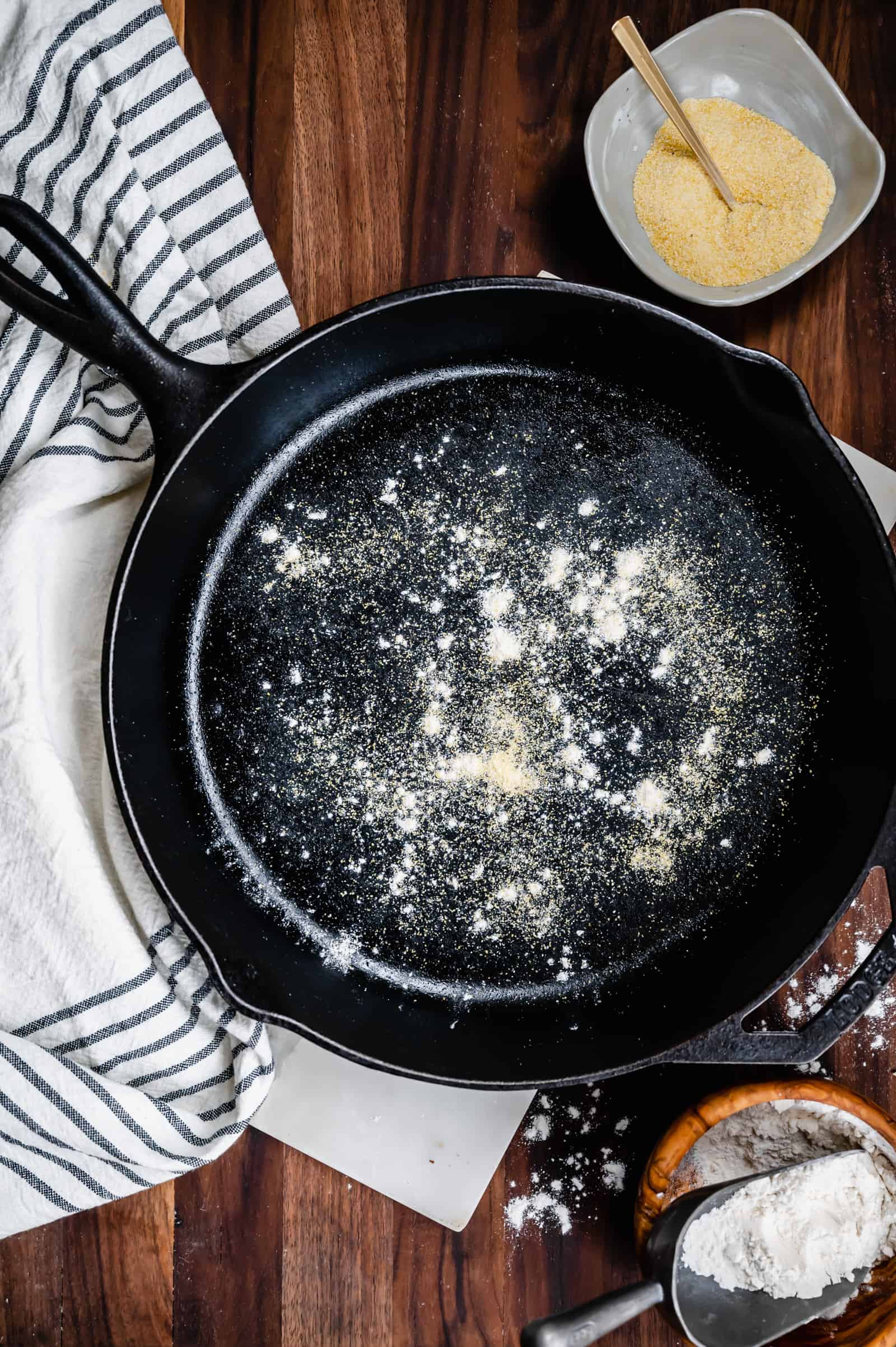 cast iron skillet with cornmeal and flour sprinkled in it with small bowls of cornmeal and flour on a walnut board.