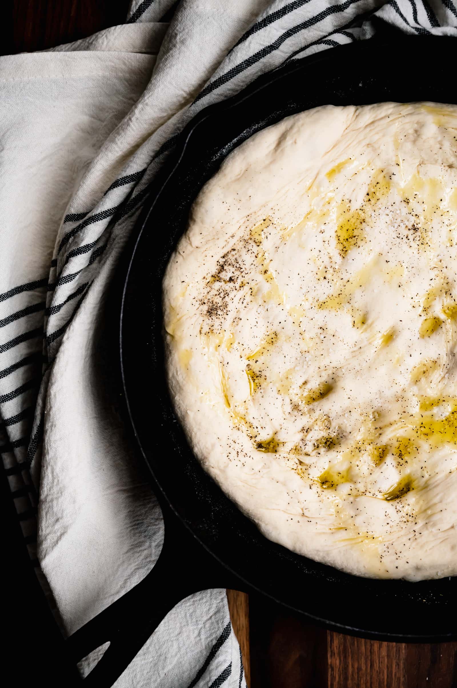 cast iron skillet with olive oil covered dough and a black and white towel.
