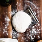 pizza dough on a floured walnut board with a scraper, a white and black towel and olive oil bottle.