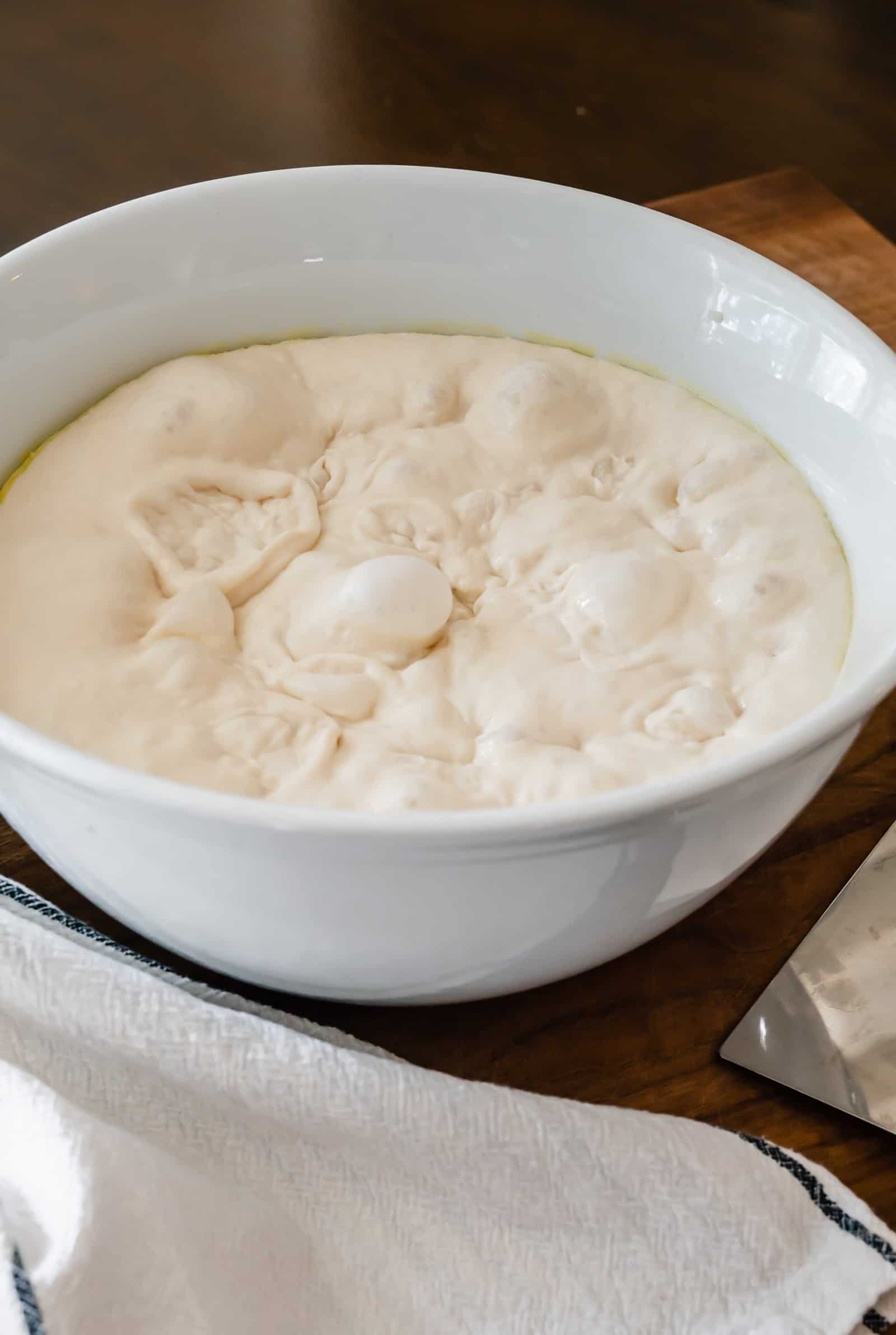 white bowl of pizza dough on walnut board with white towel