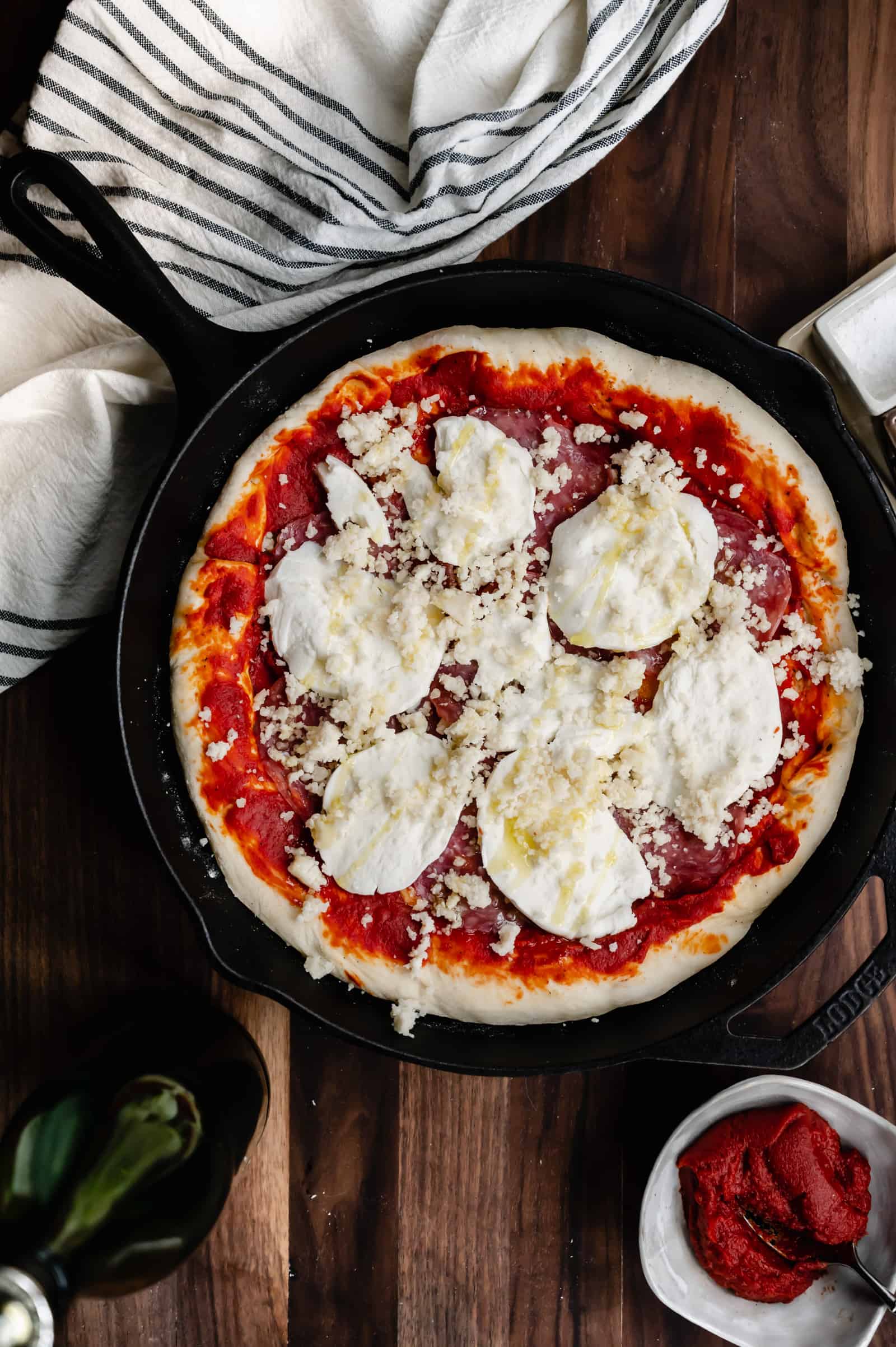 cast iron skillet pizza with soppressata, fresh mozzarella and parmesan on a walnut board with toppings and a black and white cloth.