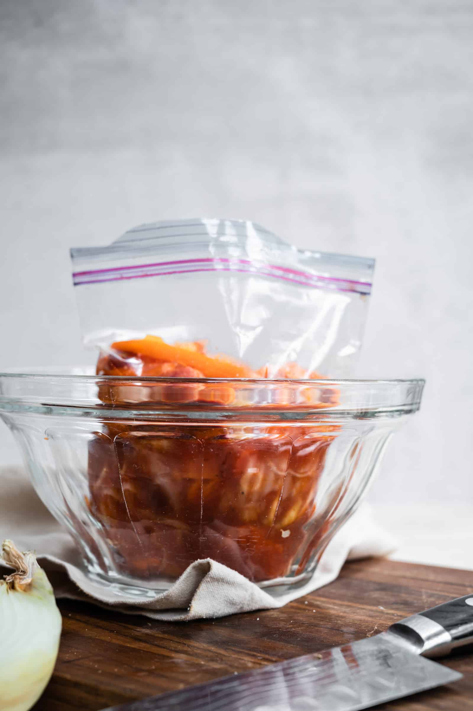 carrot salad in a ziploc bag in clear bowl on a wooden cutting board with a knife and onion.