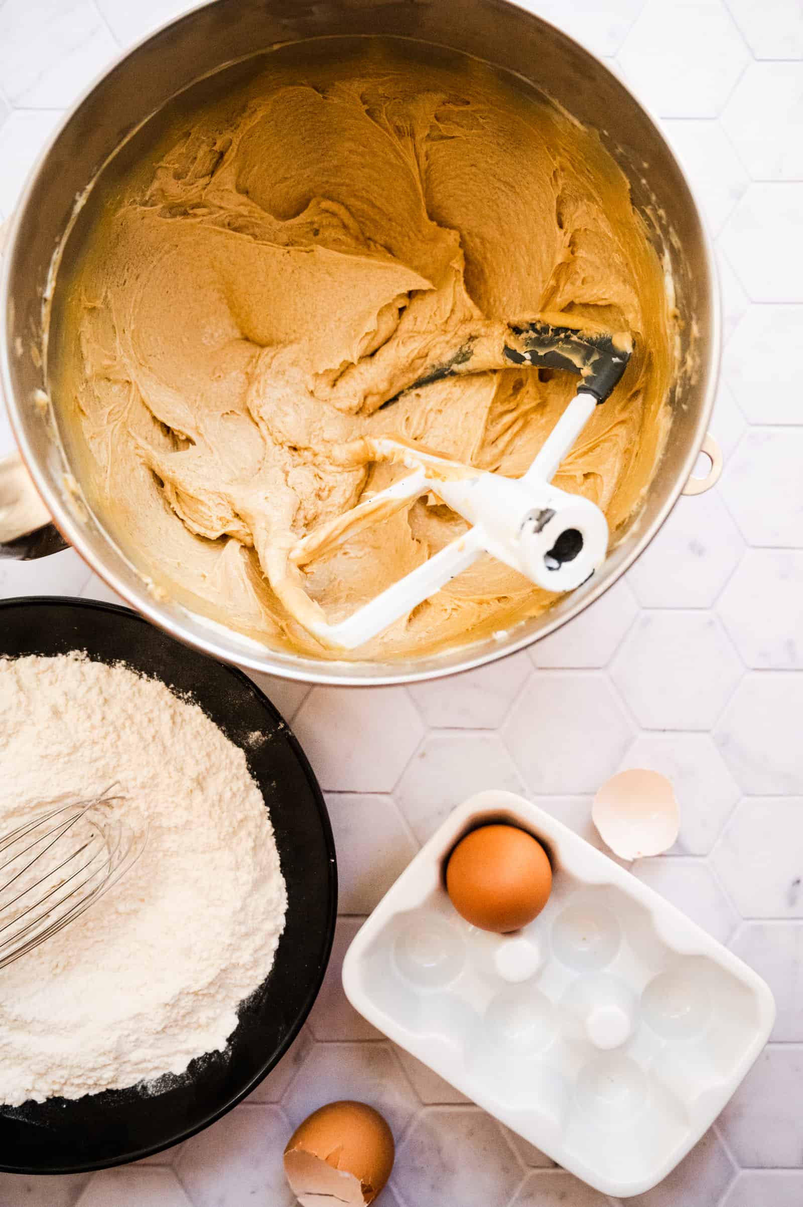 mixing bowl of cookie dough and black bowl of flour and some eggs on a marble background.