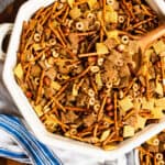 white bowl of chex mix and a wooden spoon with a blue dish towel on a wooden board.