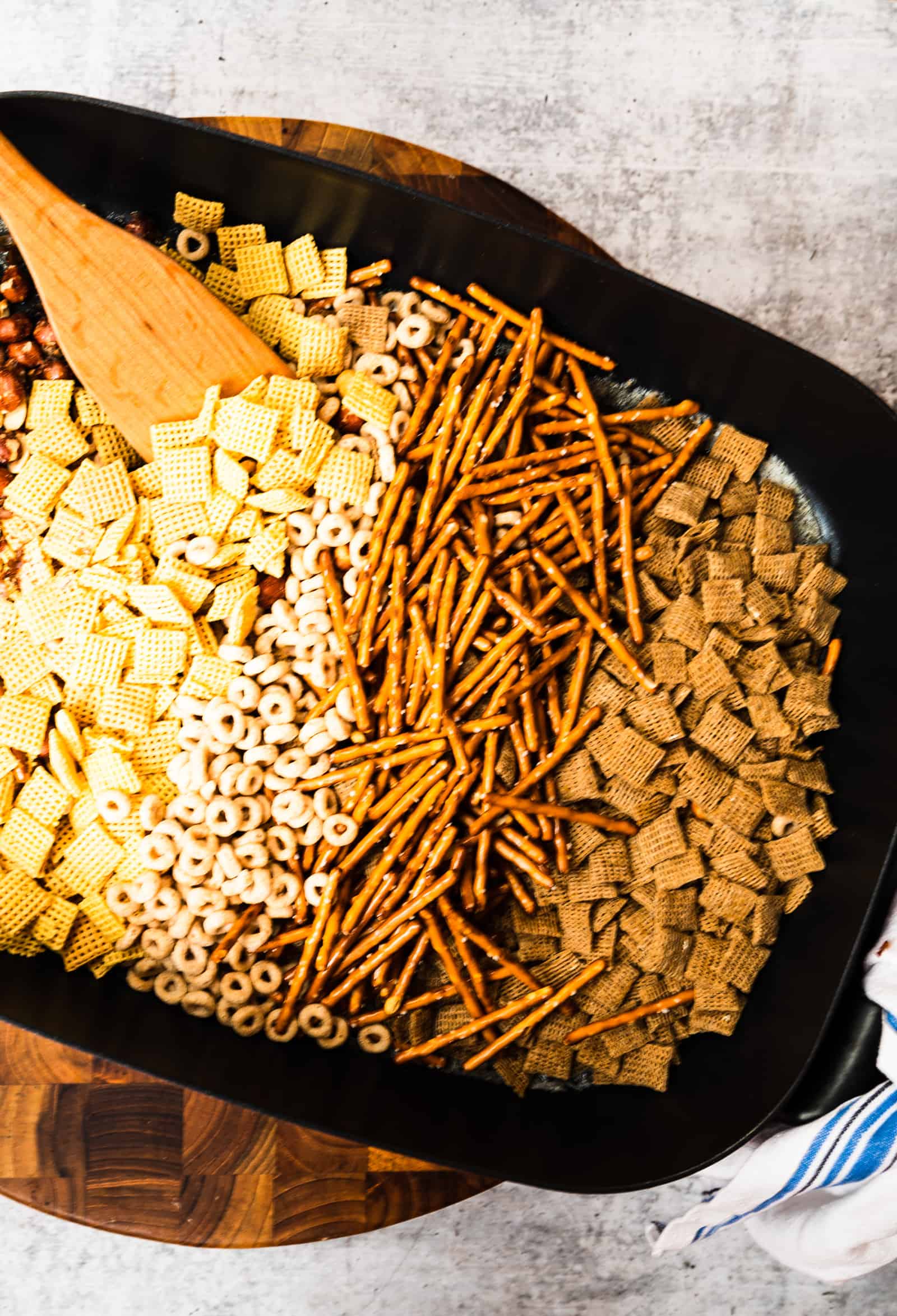 skillet of chex mix ingredients on a wooden board and blue and white towel.