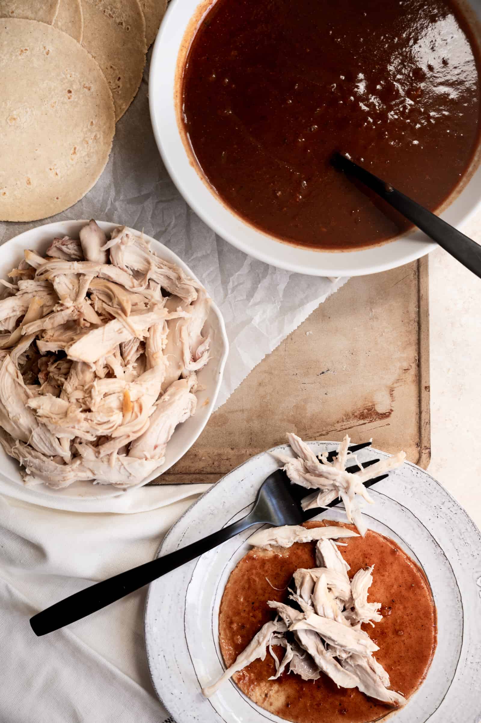 bowls of shredded chicken, enchilada sauce and a tortilla with sauce and chicken.