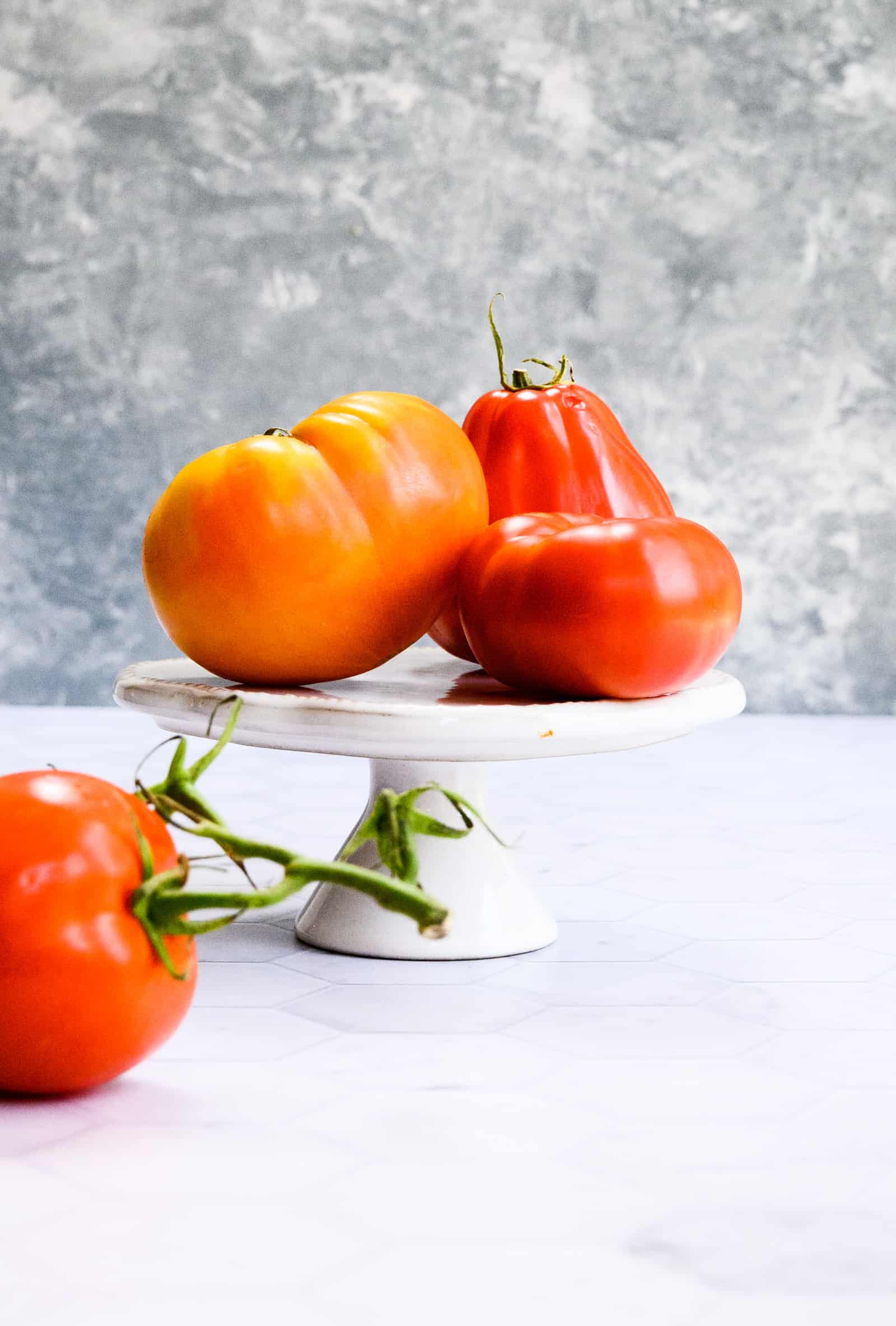 a white pedestal with three heirloom tomatoes and one tomato in the foreground.