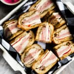 a white ceramic basket with black and white gingham napkin in it filled with pressed meat and cheese sandwiches.