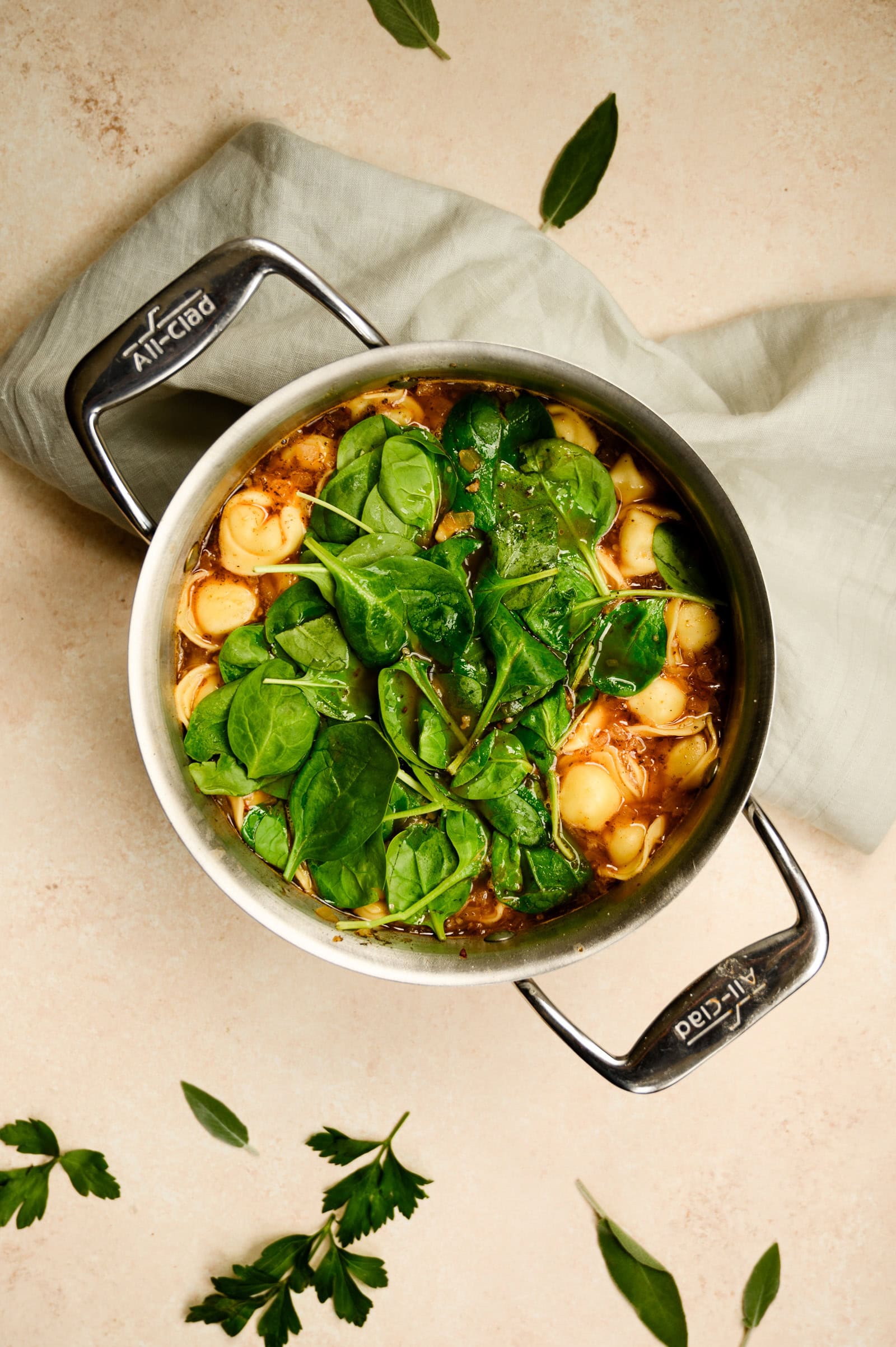 soup pan of turkey tortellini soup sits on a light surface, featuring tortellini, spinach leaves, and broth.