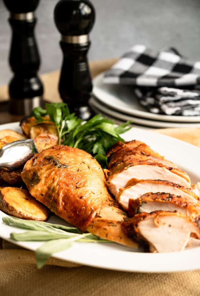 white platter of sliced roast turkey breast with the skin on it and some sage and parsley.