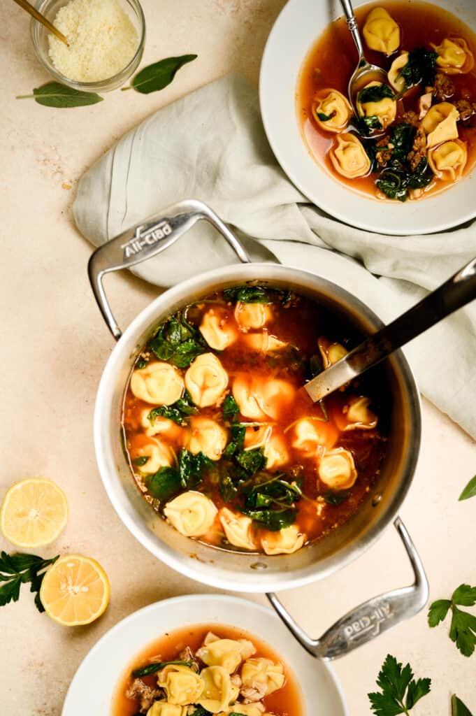 stockpot of tortellini soup with two white serving bowls of soup with spoons in them.