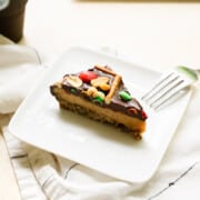 triangle piece of peanut butter pie on a white plate with ganache, pretzels and candy on top