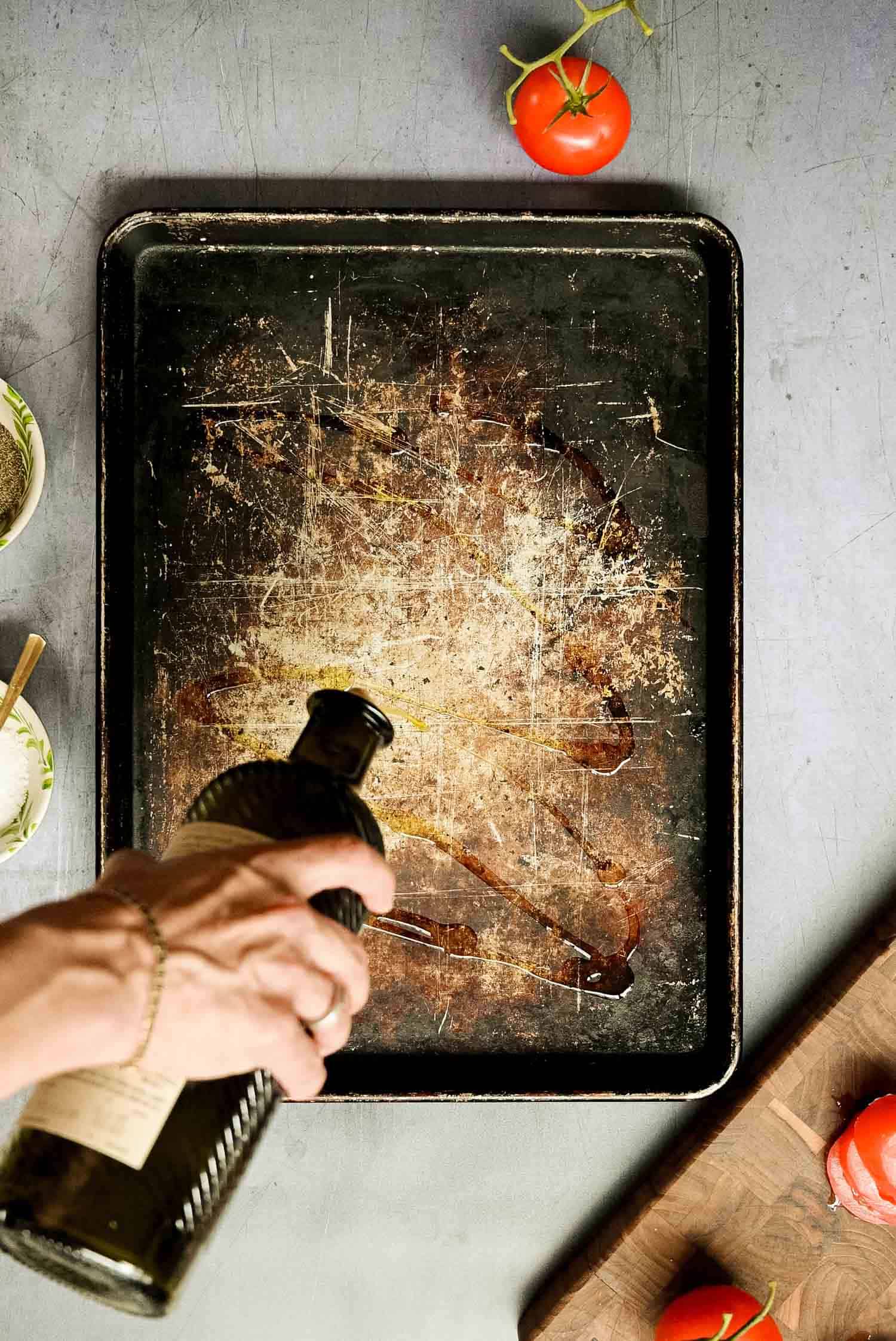 hand pouring oil on a sheet pan with some tomatoes nearby