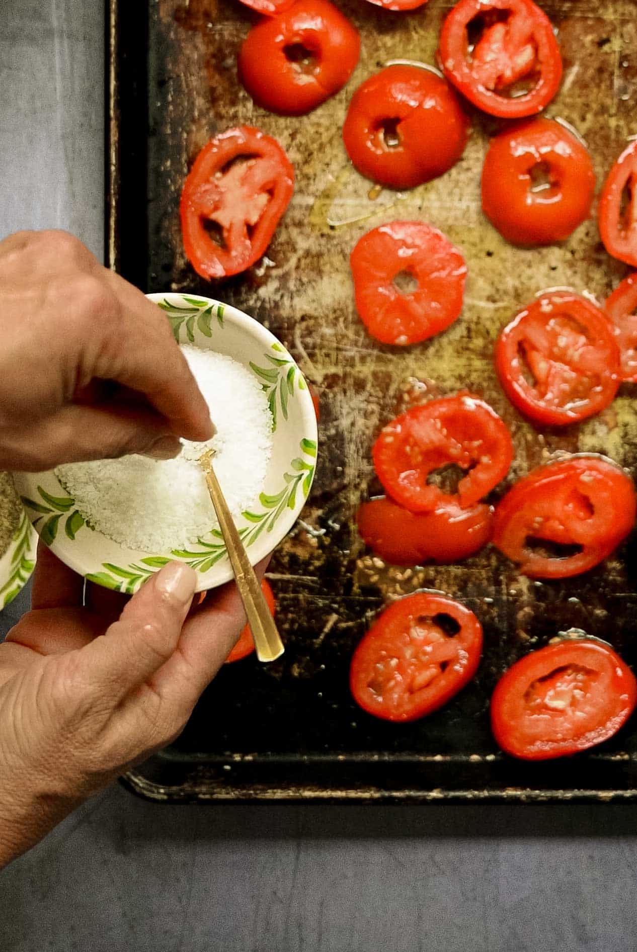 hand grabbing salt out of a white and green bowl over some tomatoes