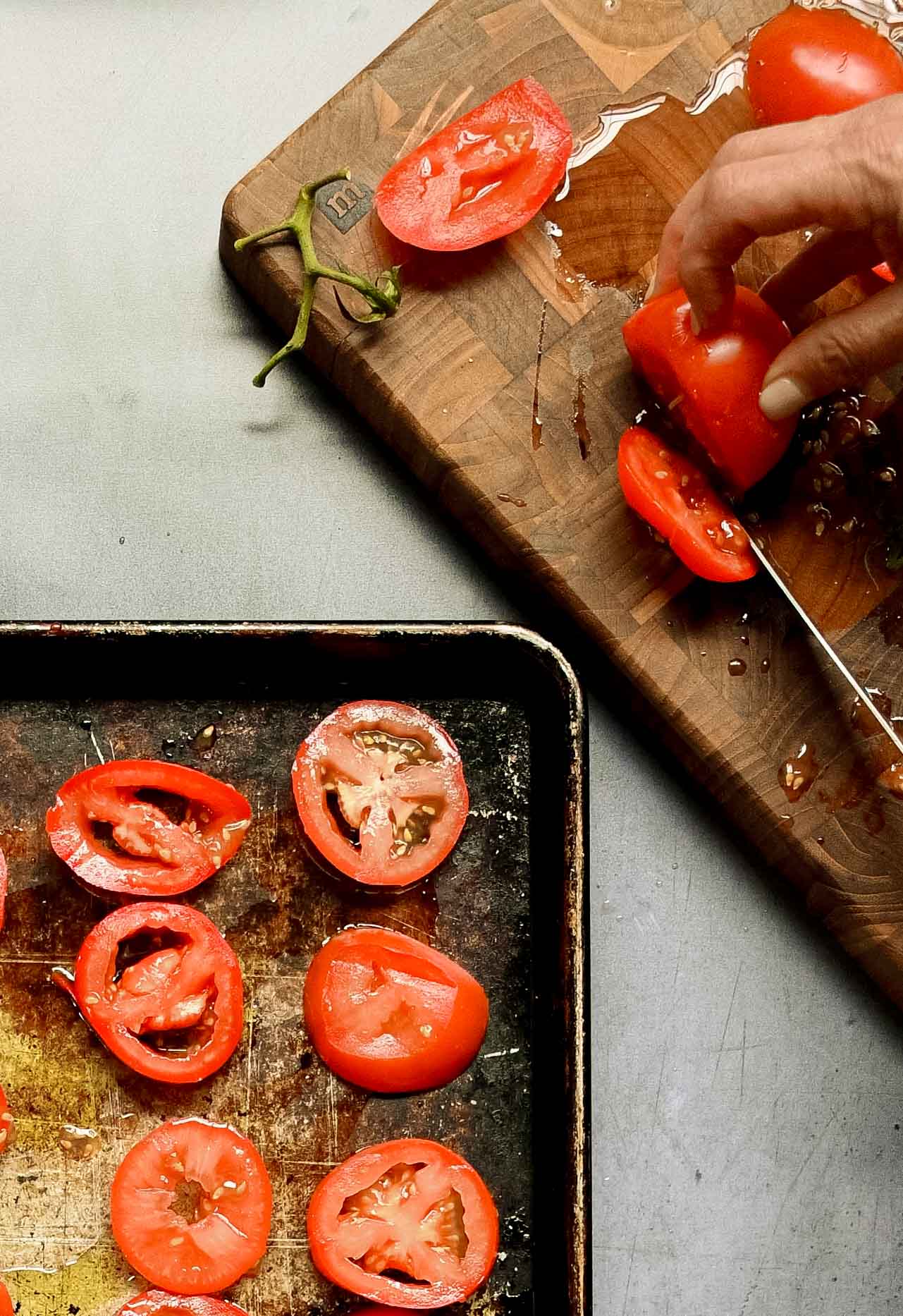 hand slicing some tomatoes on a wooden cutting board near some on a sheet pan