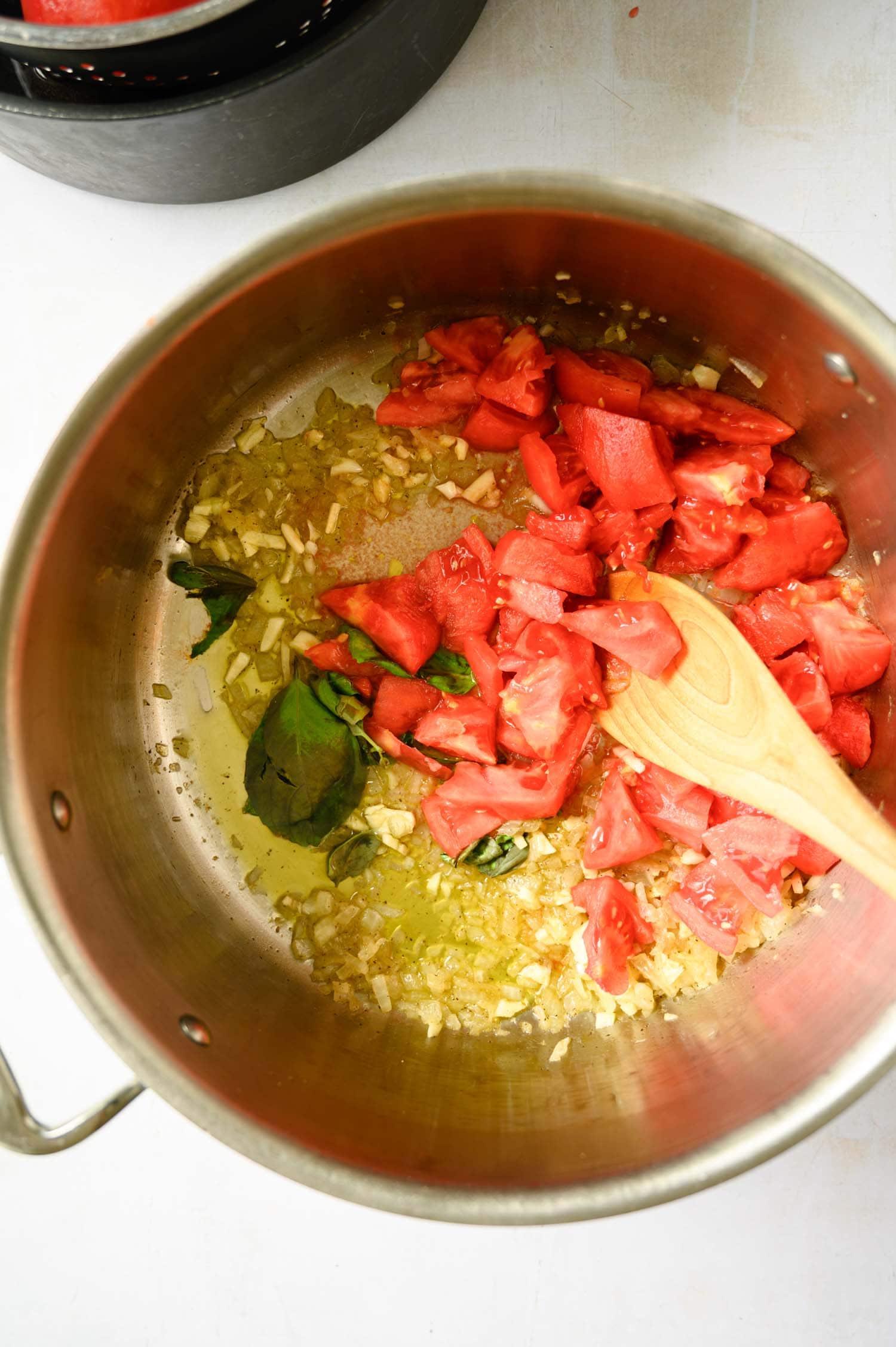 Top-down view of a metal pot with chopped tomatoes, onions, and basil leaves in olive oil for marinara.