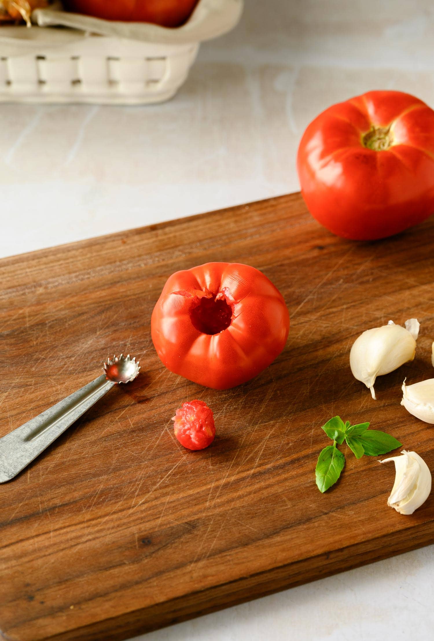 A tomato with its core removed sits on a wooden cutting board next to a small scoop, a piece of tomato core, fresh basil leaves, and three garlic cloves.