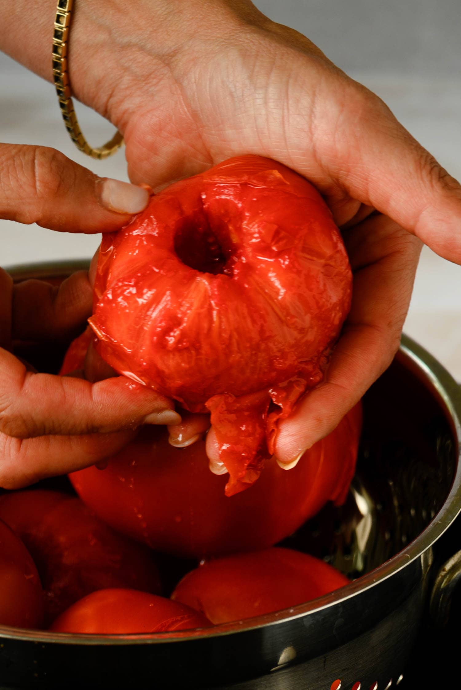 Hands peeling a blanched tomato over a bowl filled with other peeled tomatoes, each destined for a fresh marinara sauce.
