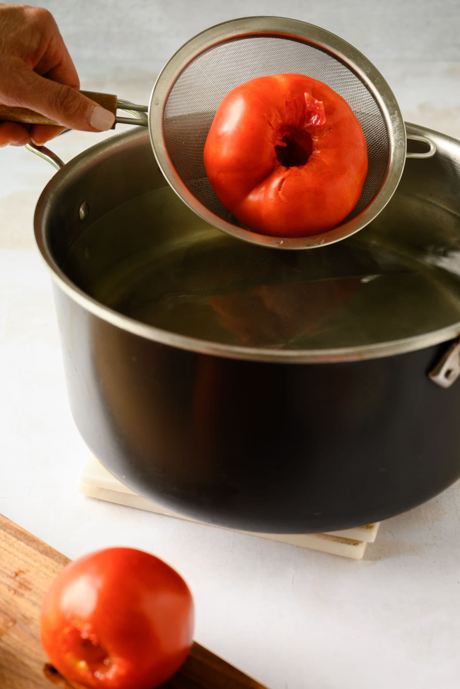 A hand holds a metal strainer with a tomato over a pot of hot water, to peel the tomato for tomato sauce.