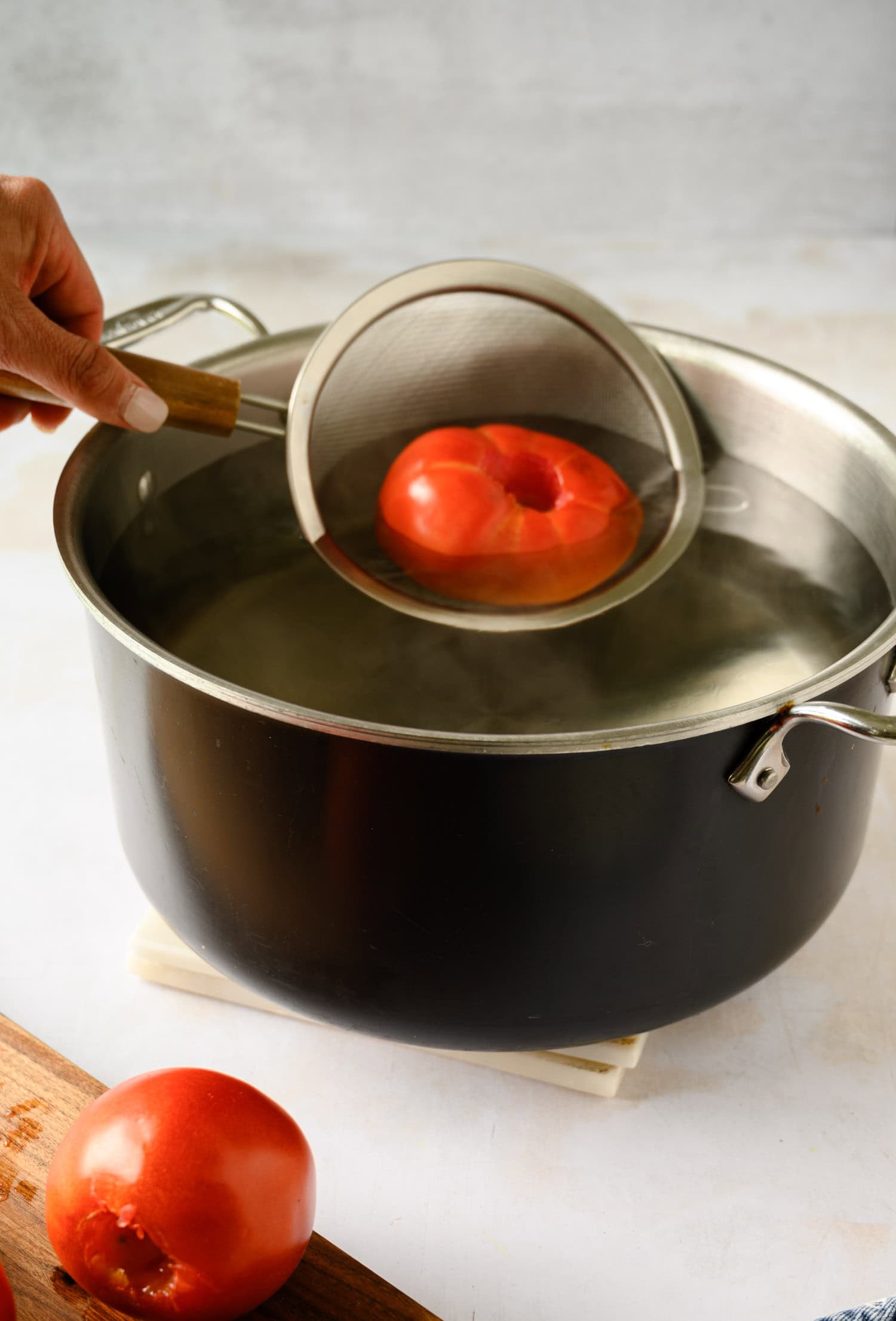 A person uses a mesh strainer to place a tomato into a pot of boiling water on the stove, preparing the base for fresh marinara sauce.
