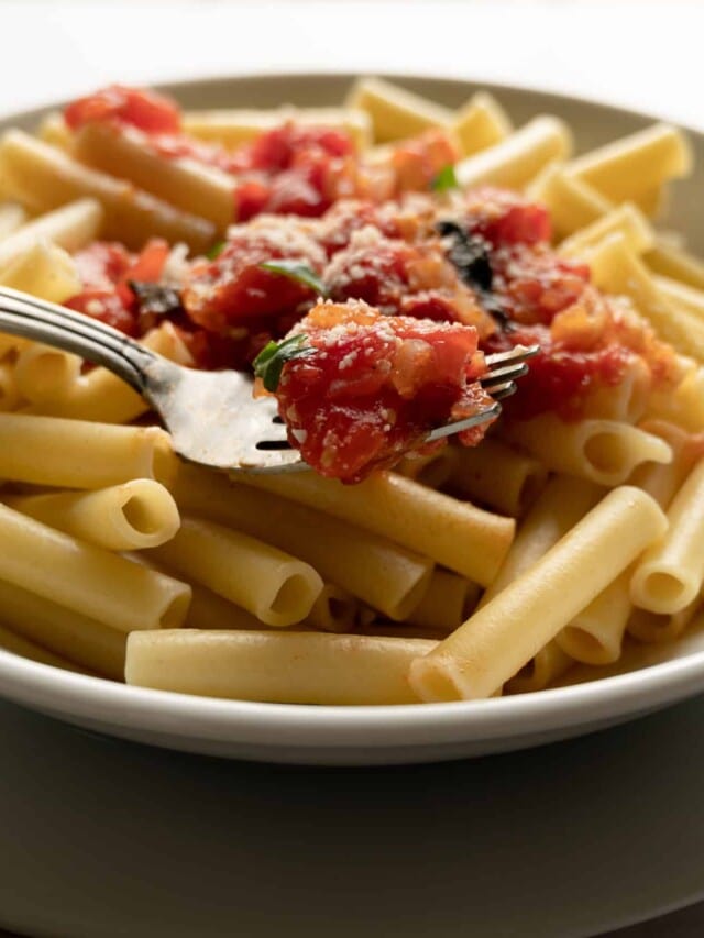 white bowl of ziti pasta with some fresh marinara sauce and a fork in front of a window