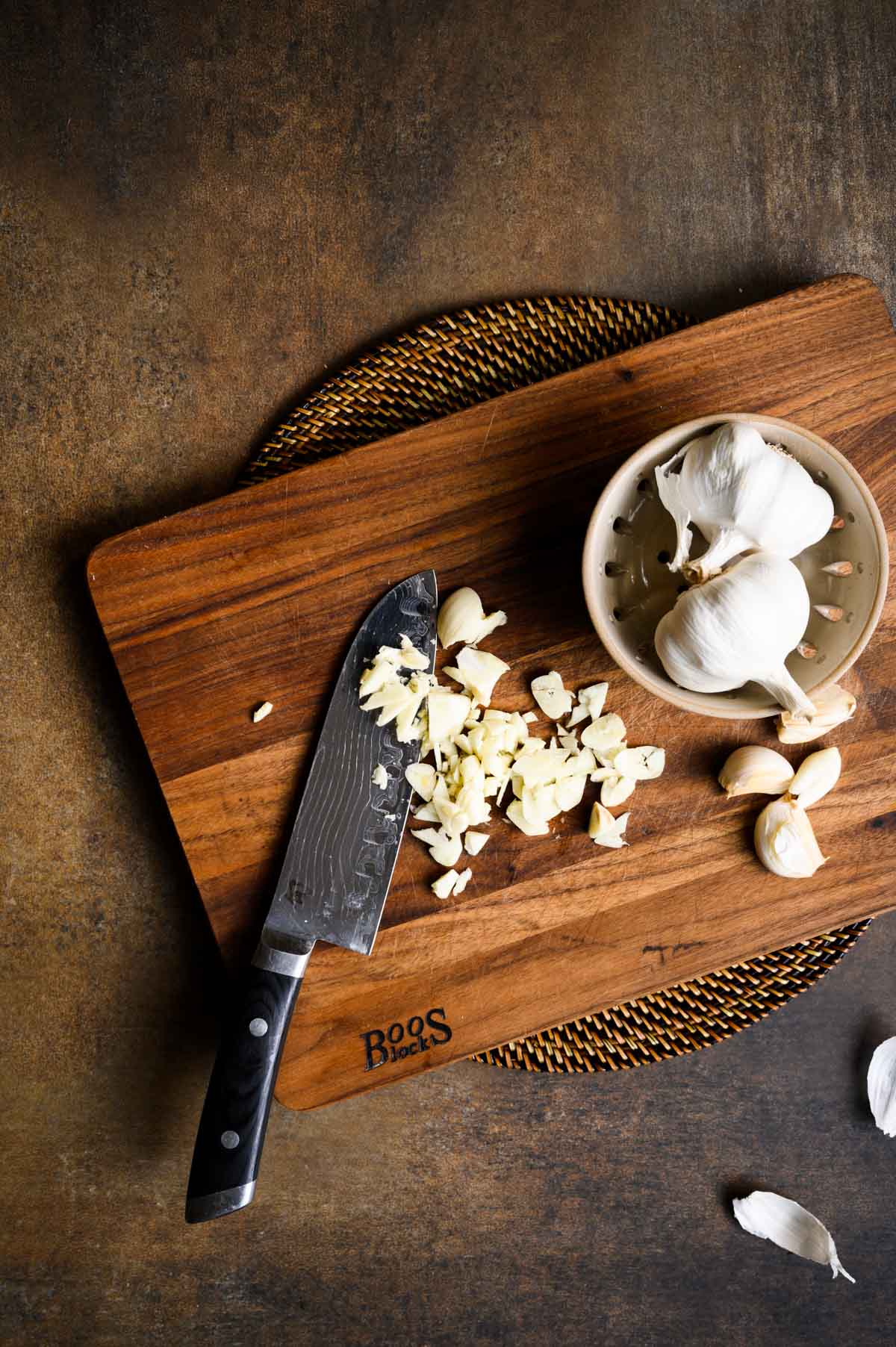 Chopped garlic and a knife on a wooden cutting board, with whole garlic bulbs in a small bowl on the side—perfect ingredients for making classic spaghetti with garlic and oil.