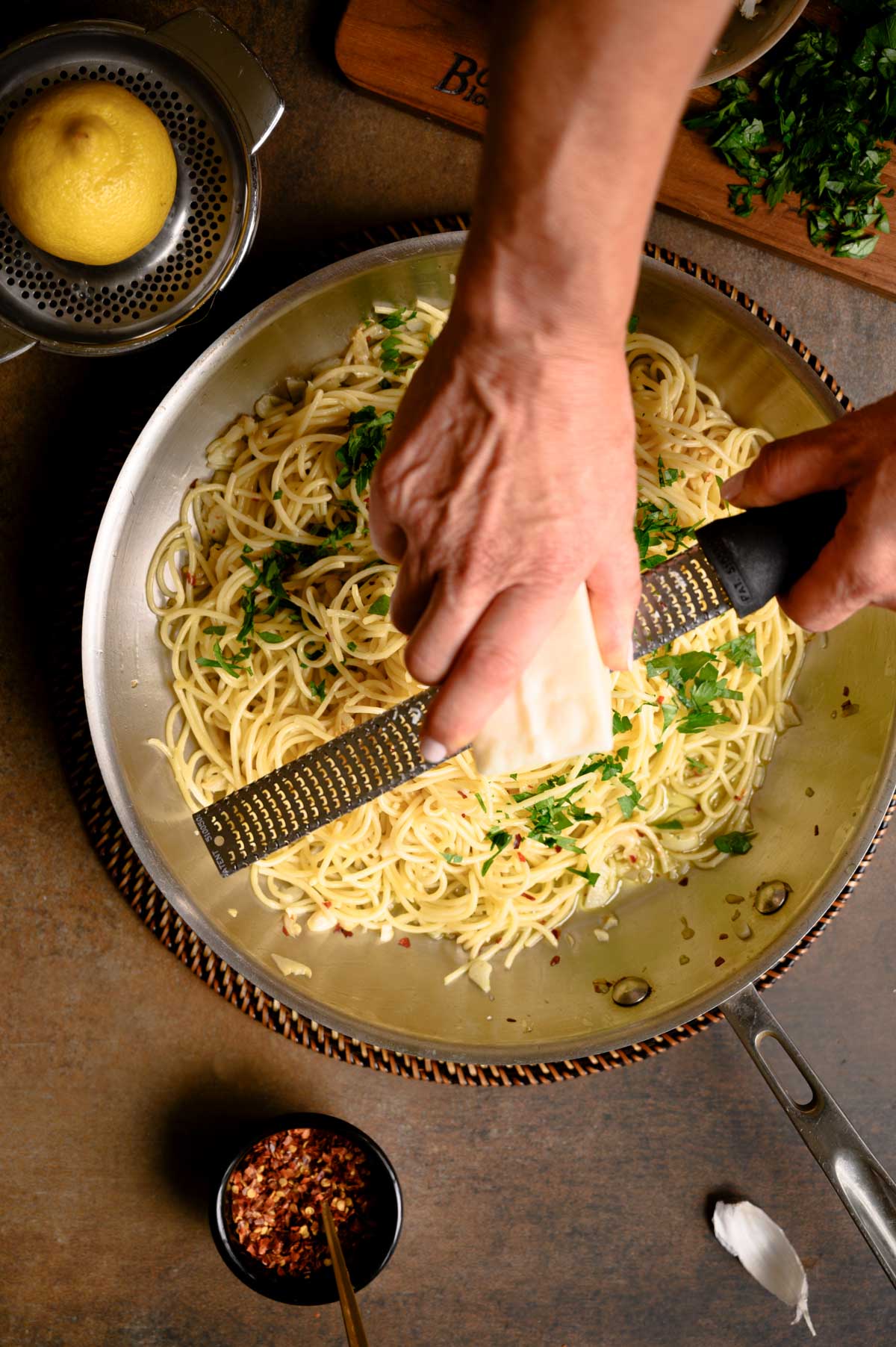 A person grates cheese over a pan of spaghetti with garlic and oil, garnished with herbs, while lemon, parsley, garlic, and red pepper flakes are scattered on the surrounding table.