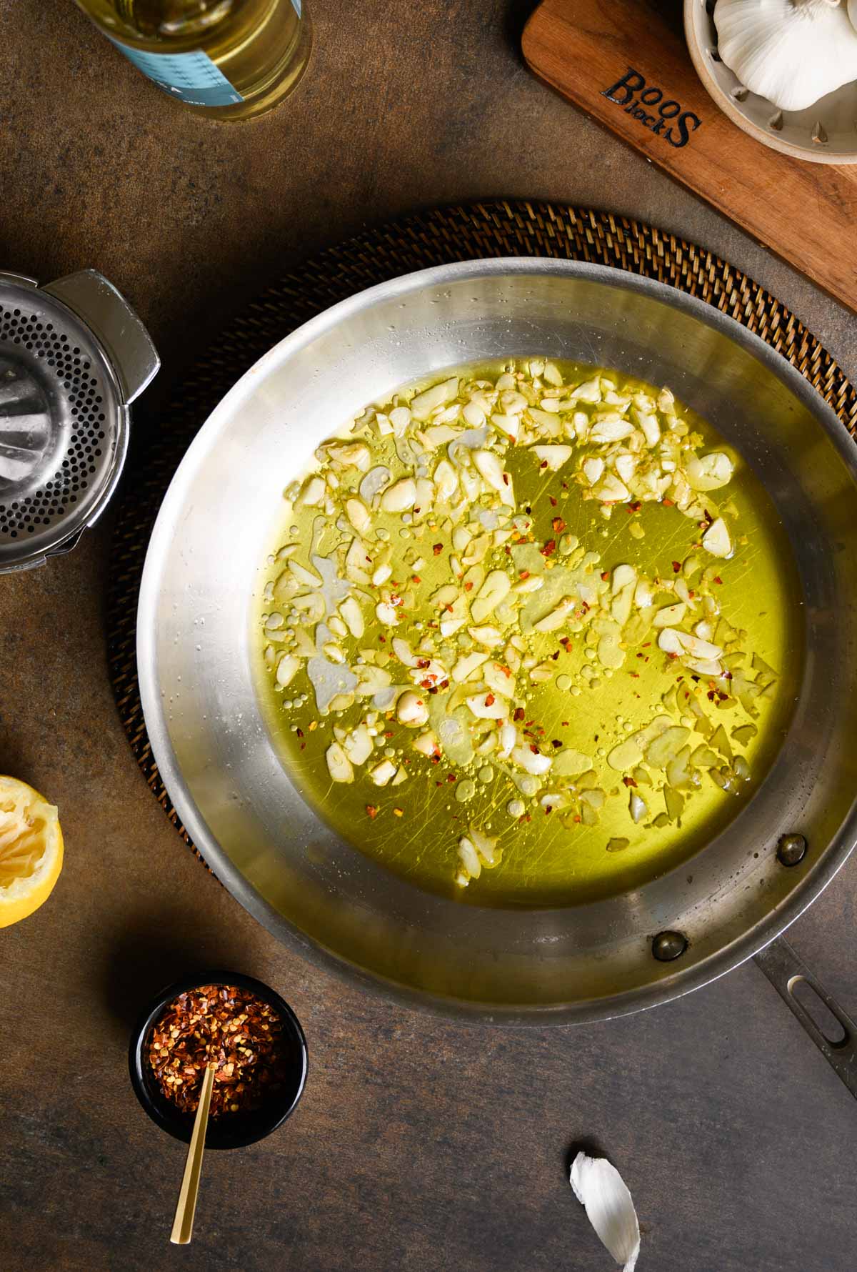 A stainless steel pan filled with olive oil, sliced garlic, and red pepper flakes—perfect for making spaghetti with garlic and oil—sits on a dark countertop surrounded by various cooking ingredients.