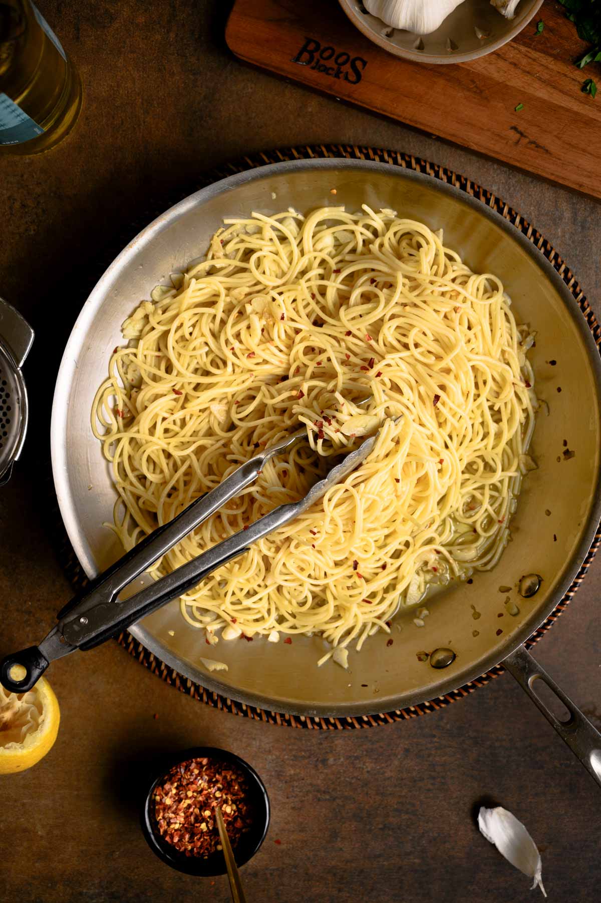 A pan of spaghetti with garlic and oil, sprinkled with red pepper flakes, sits on a brown surface. Nearby are a lemon wedge, minced garlic, and a small bowl of chili flakes. Tongs rest in the flavorful pasta, ready to serve.