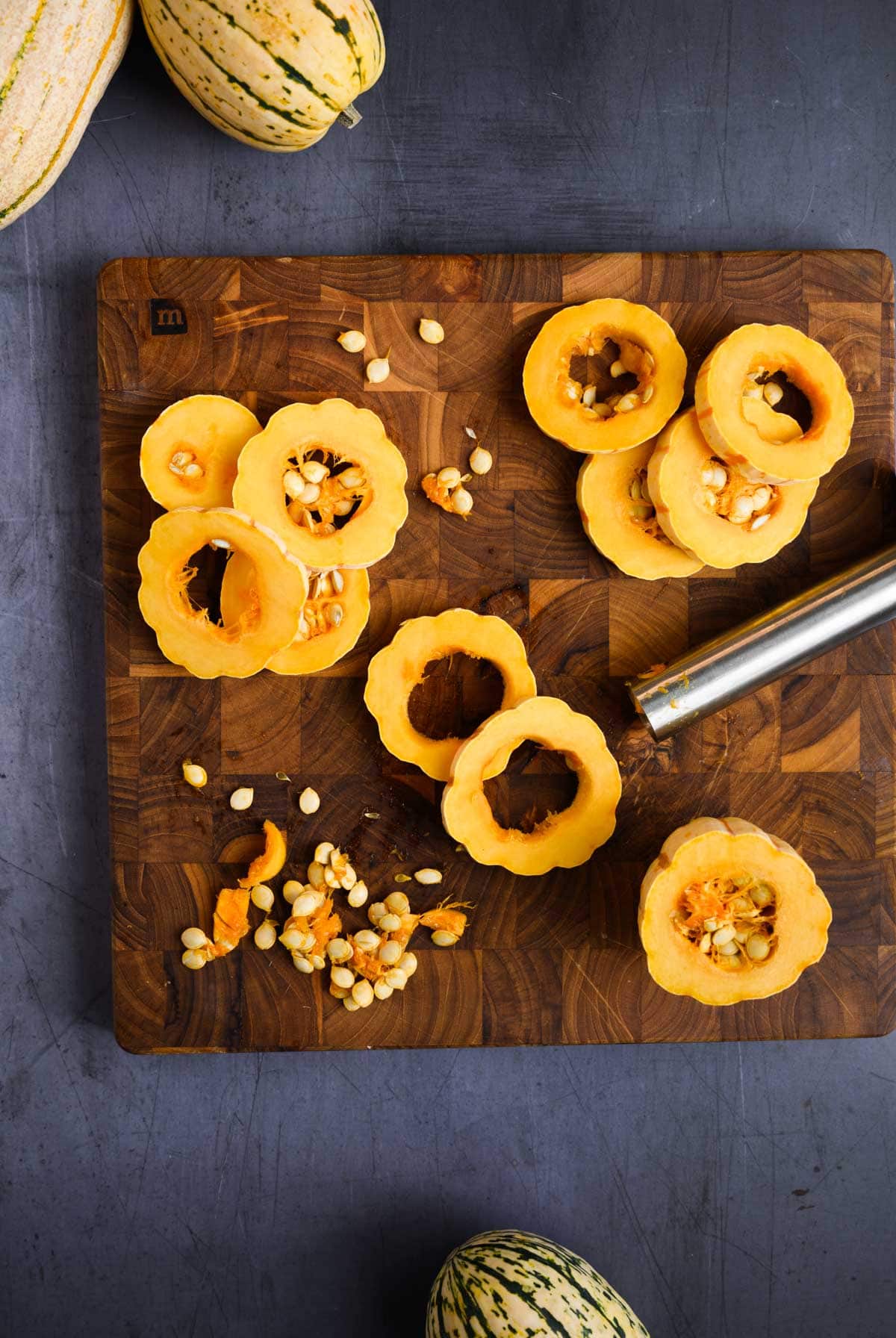 A wooden cutting board holds sliced delicata squash rings—some seeded, some cored with a metal tool—plus loose seeds.