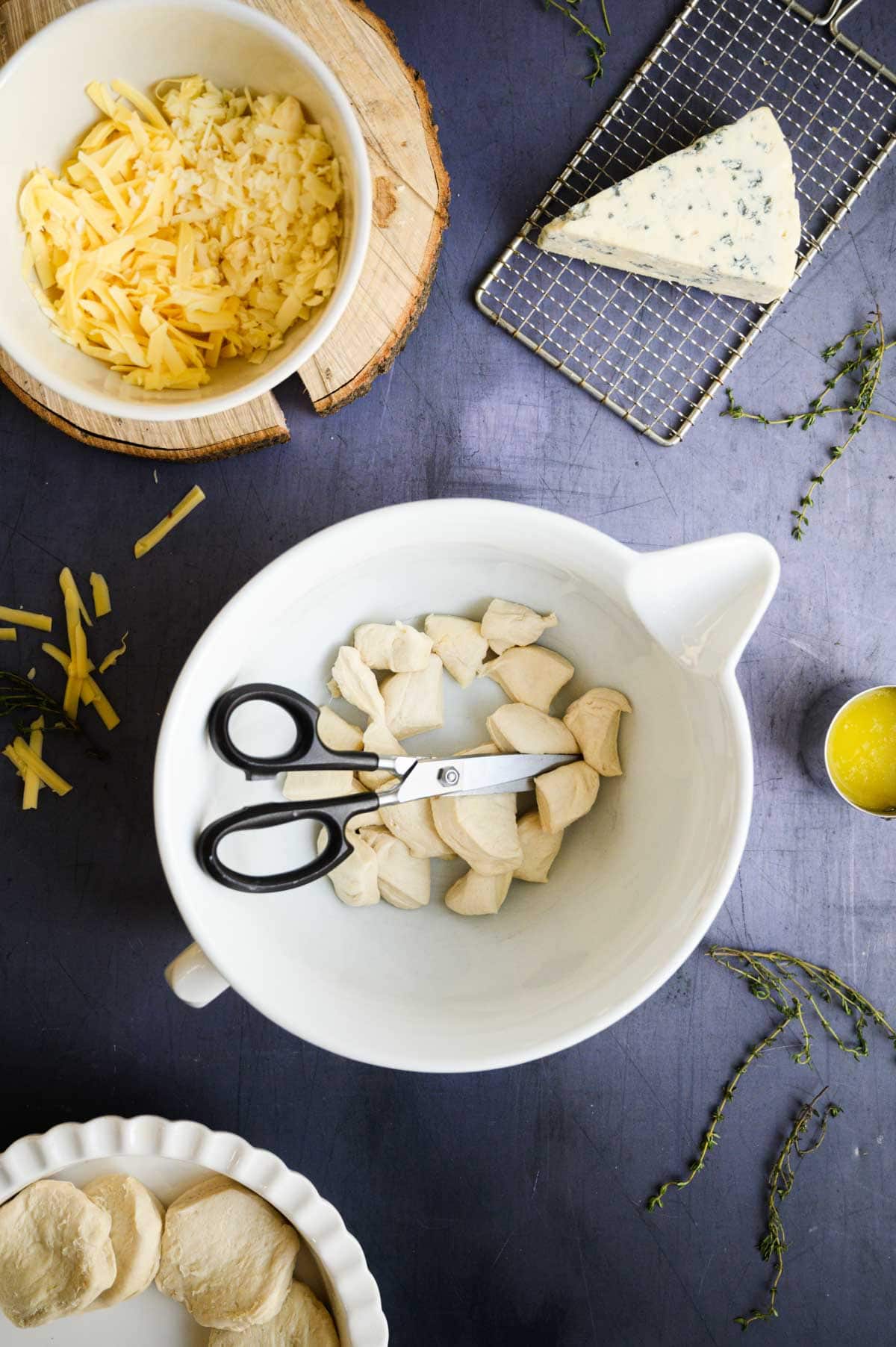 A bowl of cut dough pieces for cheesy monkey bread with kitchen shears, surrounded by shredded cheese, a wedge of blue cheese, melted butter, and fresh thyme.
