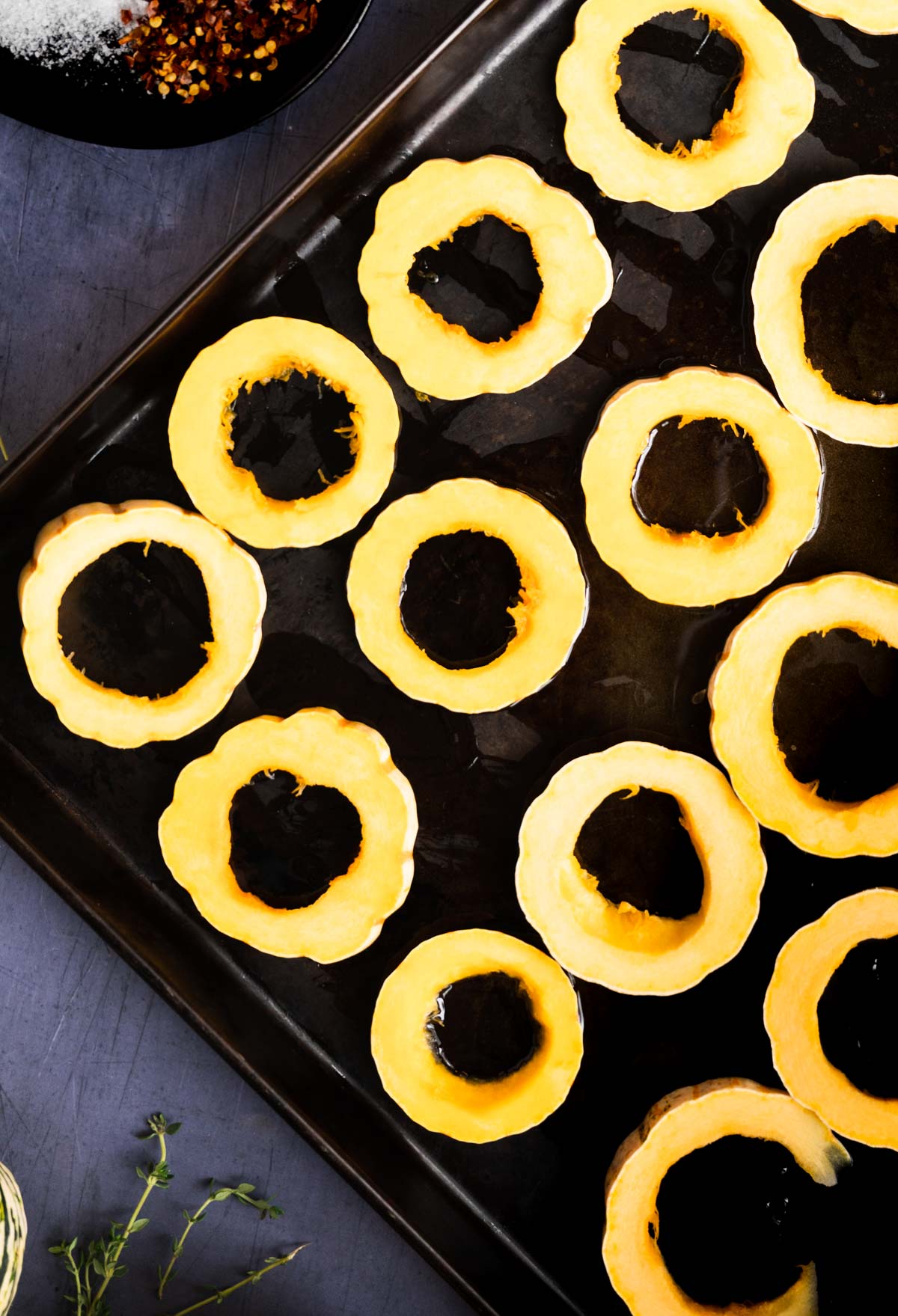 A baking sheet with evenly spaced raw acorn squash rings, lightly coated with oil, ready for roasting. Some herbs and seasoning are visible in the background, perfect inspiration for your next delicata squash recipe.