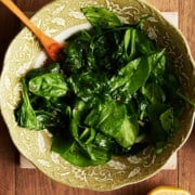 cooked spinach in a green floral bowl with a wooden fork and halved lemon and crushed red pepper.