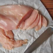 Raw chicken breast on parchment and cutting board being sliced with a knife.