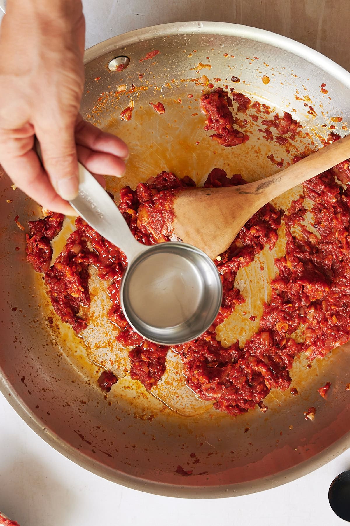A hand holds a measuring cup of vodka above a pan where tomato paste is being stirred with a wooden spoon.